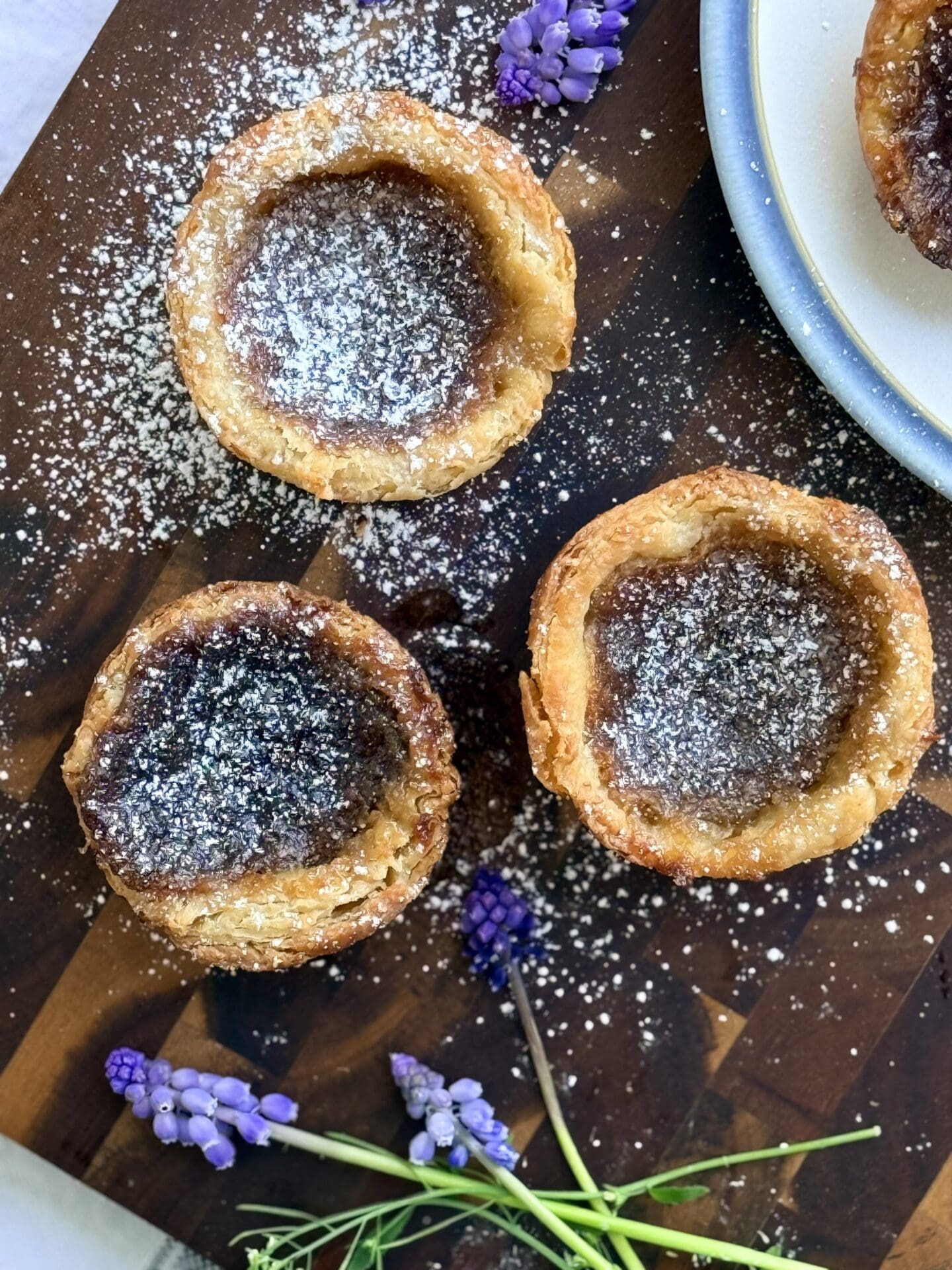 A collection of 3 freshly baked butter tarts sit on a wood board, dusted with powdered sugar and surrounded by small purple flowers.