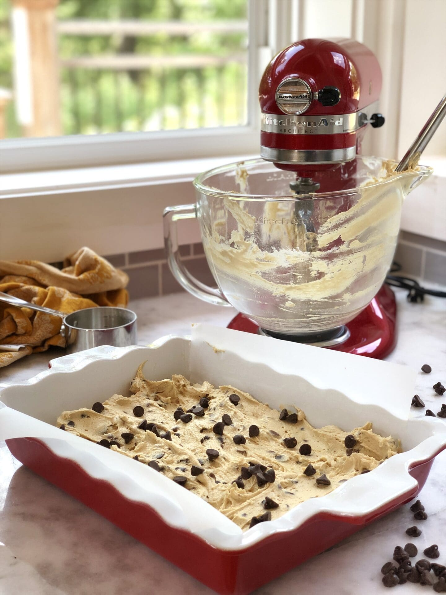 A cherry red stand mixer streaked with batter sits by a cottage window. A pan of chocolate chip blondies sits in the foreground, ready for the oven.