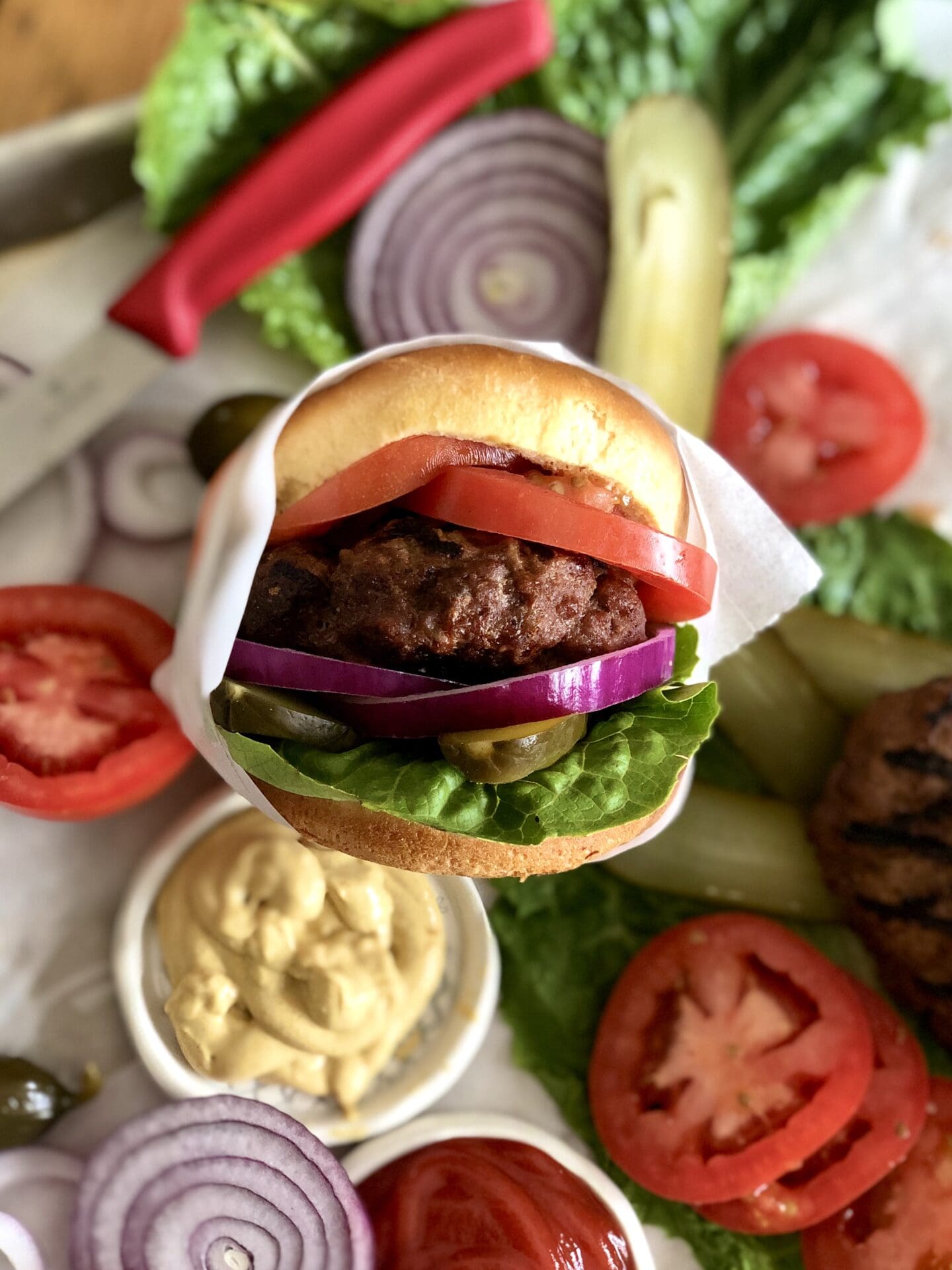 A perfect beef burger, wrapped in a golden bun and stuffed with toppings is seen in front of a tray of condiments and vegetables.