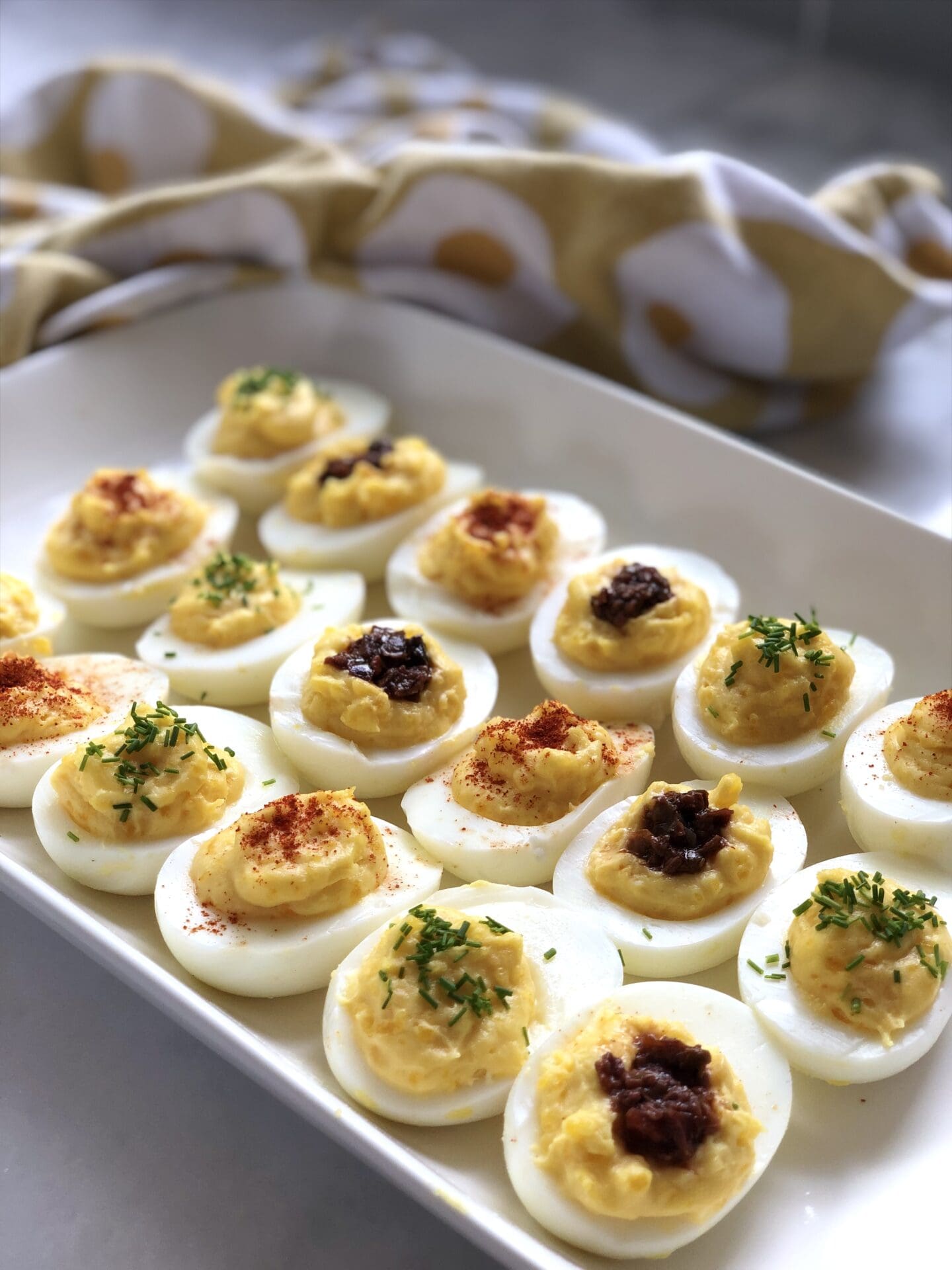Lavishly garnished devilled eggs sits on a white tray. A yellow cloth sits in the background, showing its whimsical egg pattern.