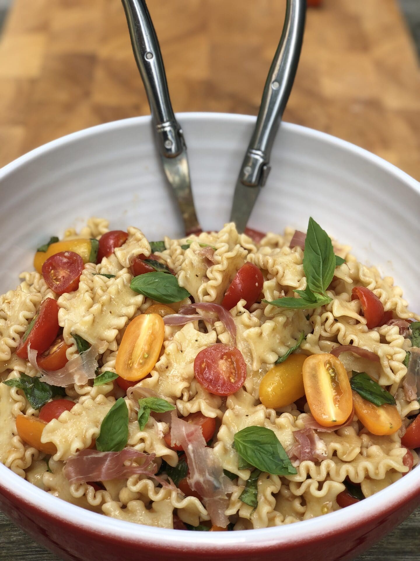 A large bowl of summer fresh pasta salad with prosciutto, cherry tomatoes and fresh basil is seen with a pair of salad servers.