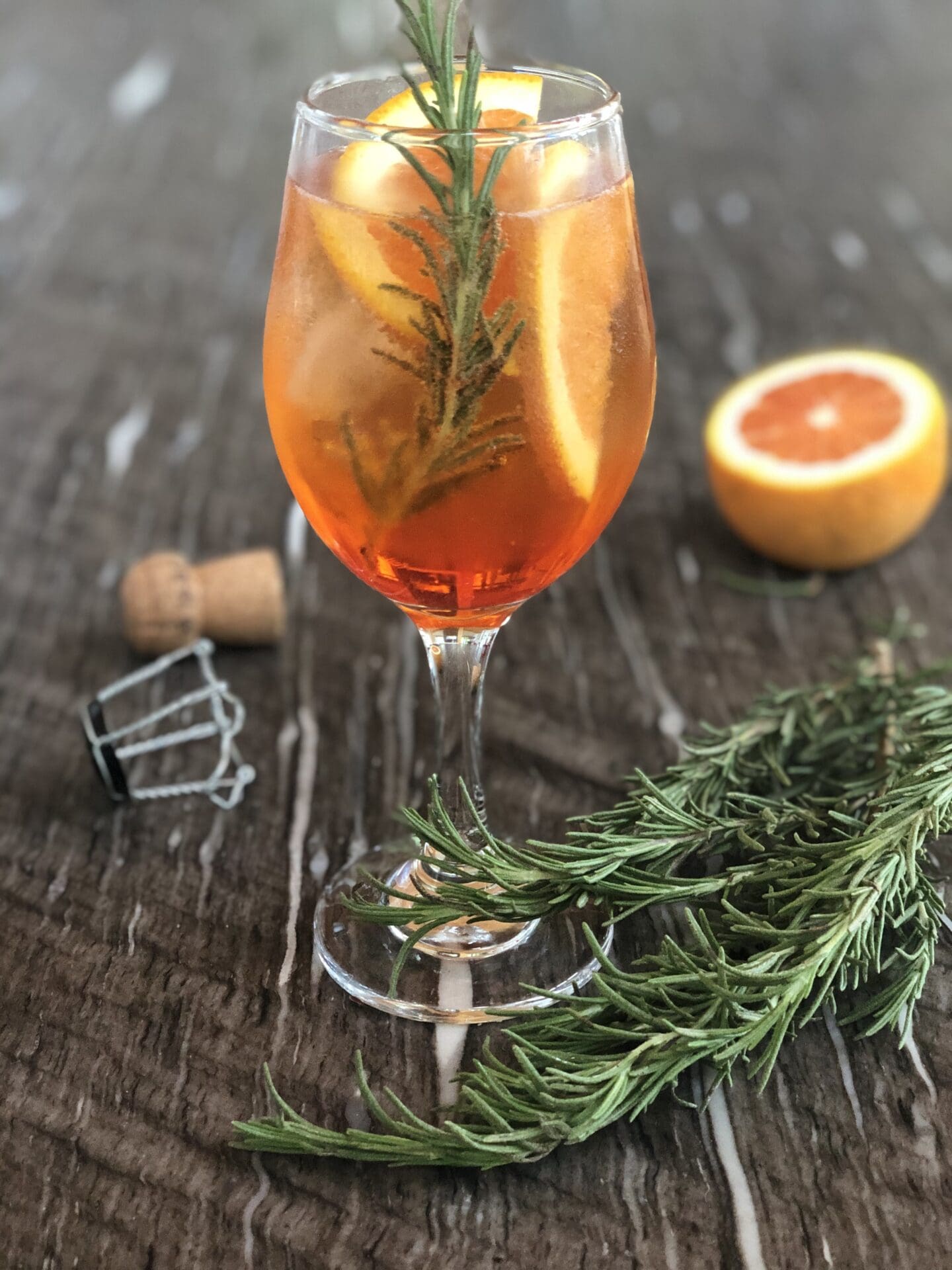An Aperol Spritz sits on a granite counter surrounded by fresh rosemary, oranges slices and the cork from the prosecco bottle.