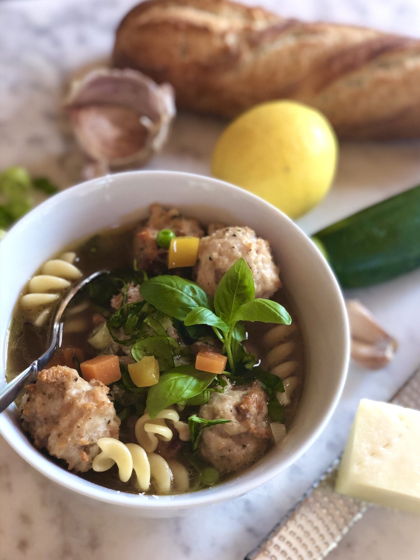 A bowl of meat ball and noodle soup is shown, garnished with fresh basil and surrounded by vegetables, a wedge of pecorino cheese and a baguette.