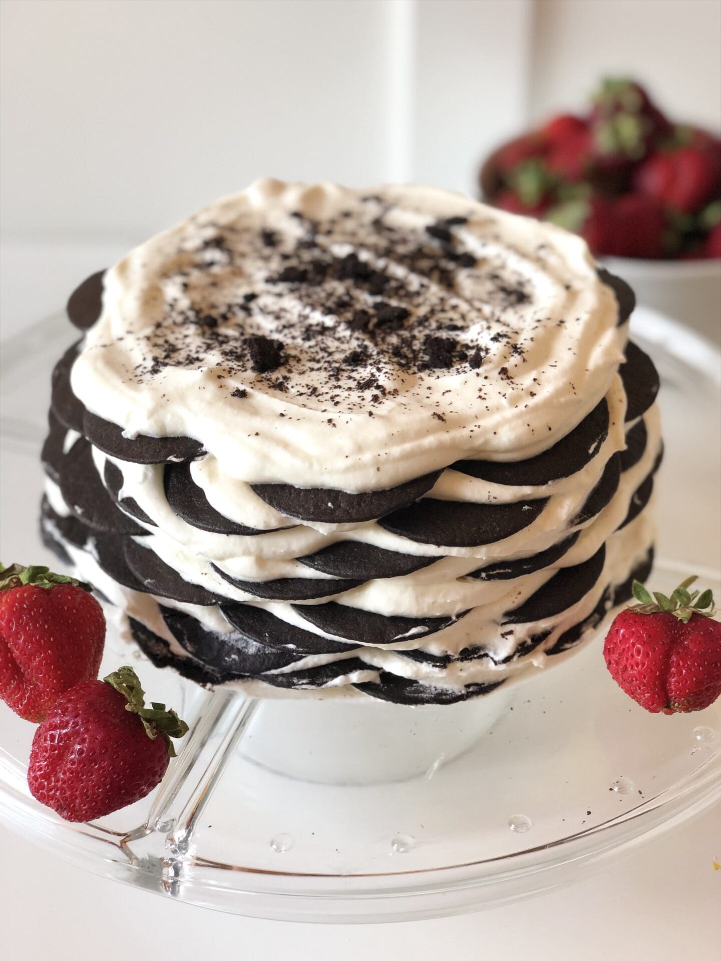 A tall ice box cake shows off its layers of chocolate cookies and whipped cream. The cake sits on a beautiful glass cake plate and is surrounded by fresh strawberries.