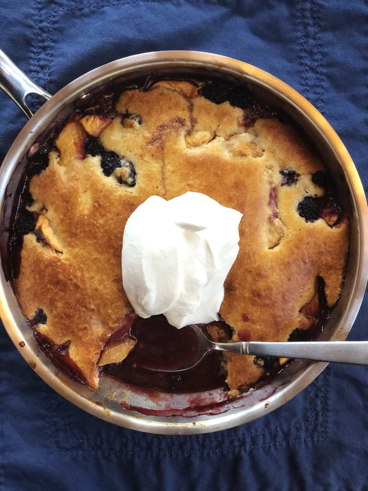 A pan of golden-topped blueberry cobbler is seen from above, topped with a generous dollop of whipped cream.