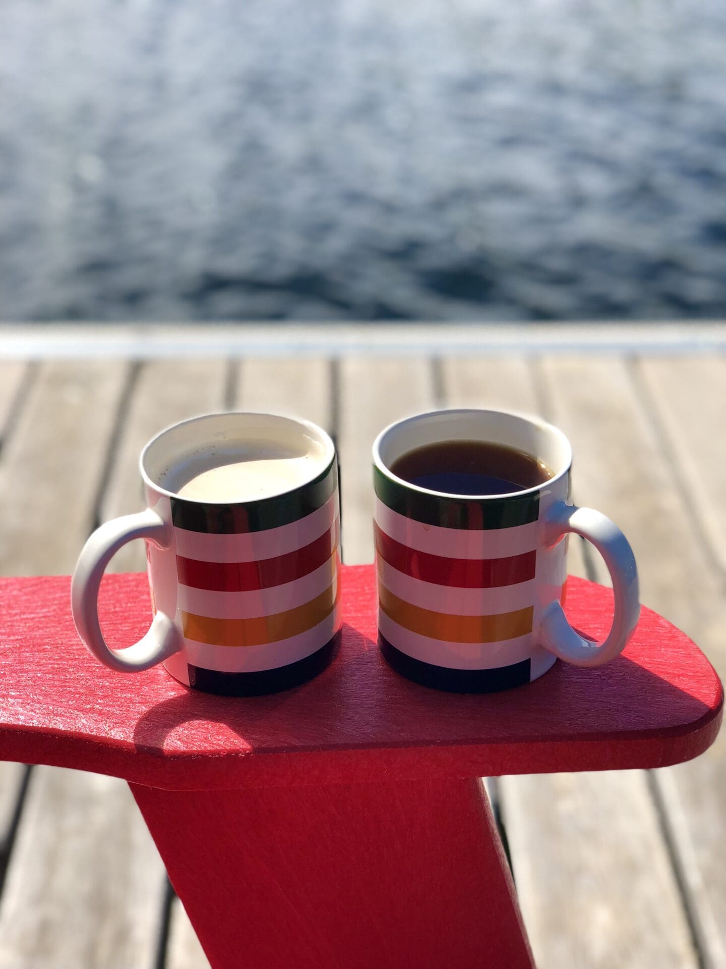 A cup of tea and a cup of coffee sit together on the arm of a red muskoka chair. The chair sits on a dock, with the blue water of the lake visible in the background.