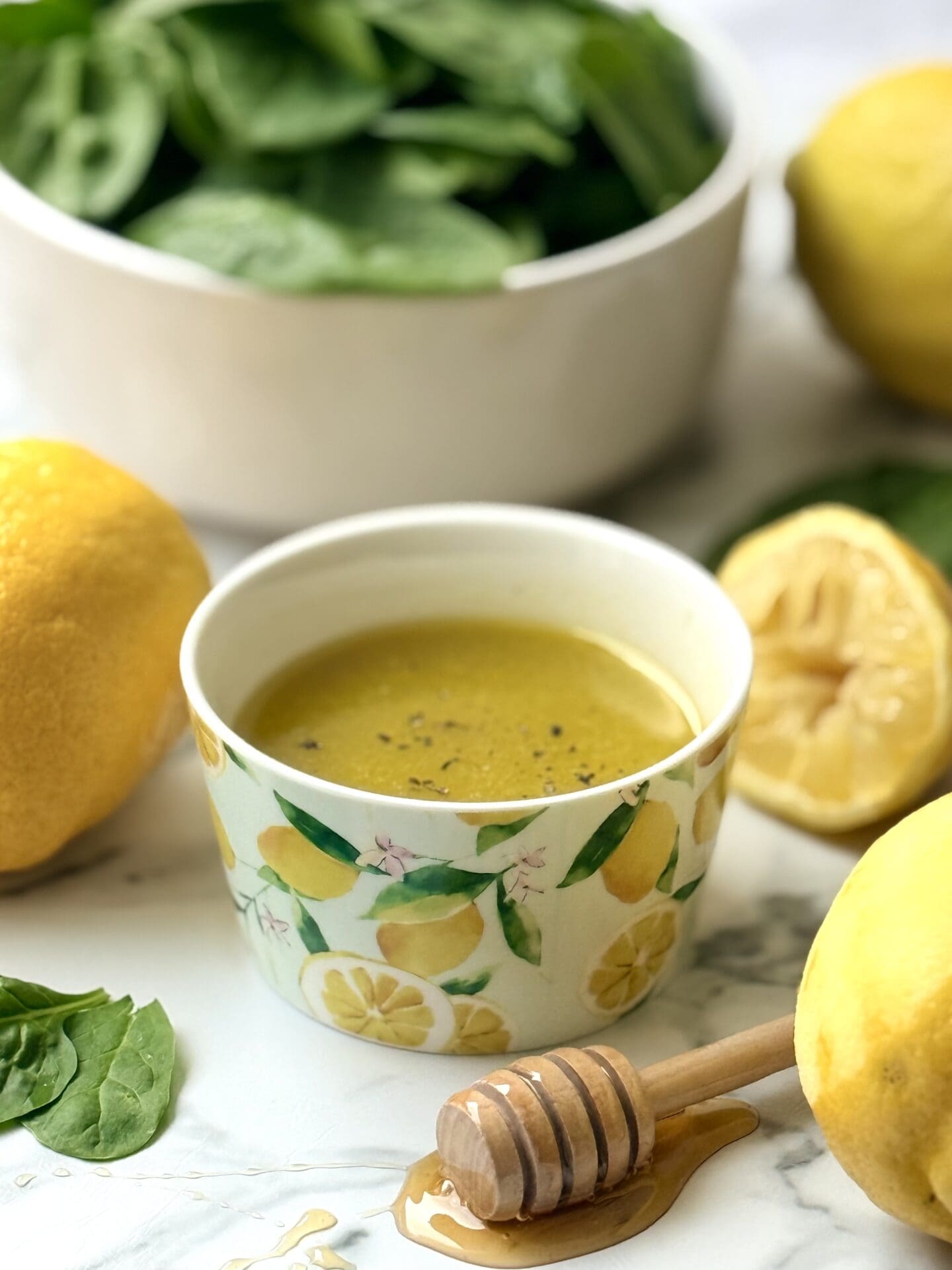 IMG_8655 A decorative bowl with a vibrant lemon print is filled with Mediterranean Lemon Vinaigrette. The bowl of dressing is surrounded by fresh lemons, honey and greens.
