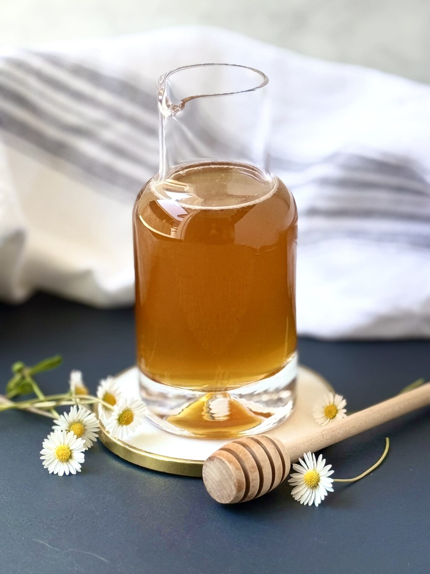 A glass bottle of honey simple syrup sits on a white and gold coaster, surrounded by tiny daisies and a honey dipper.