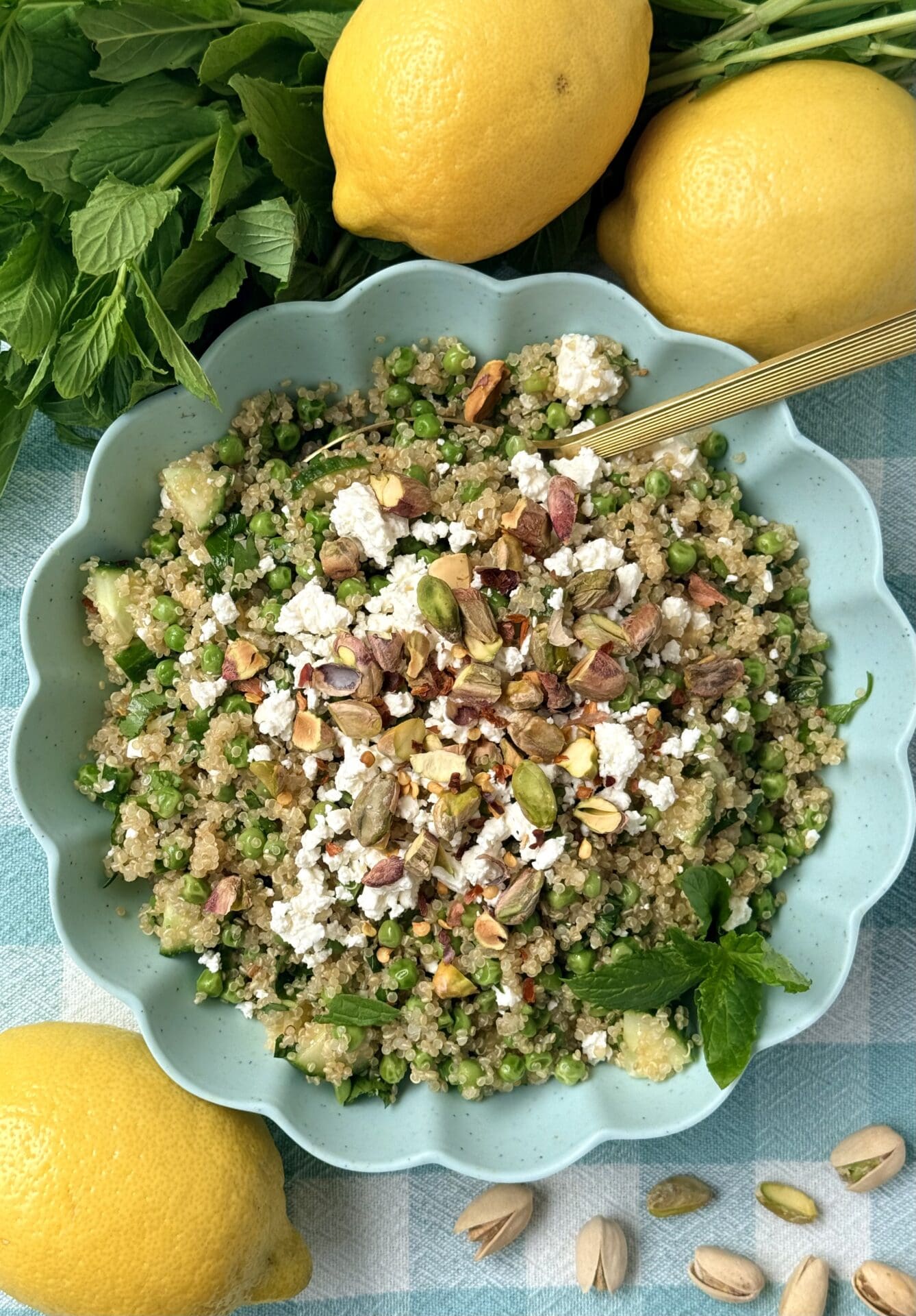 A bowl of Lemony Quinoa Salad with Spring Peas and Pistachios is seen from above, surrounded by fresh mint and lemons.