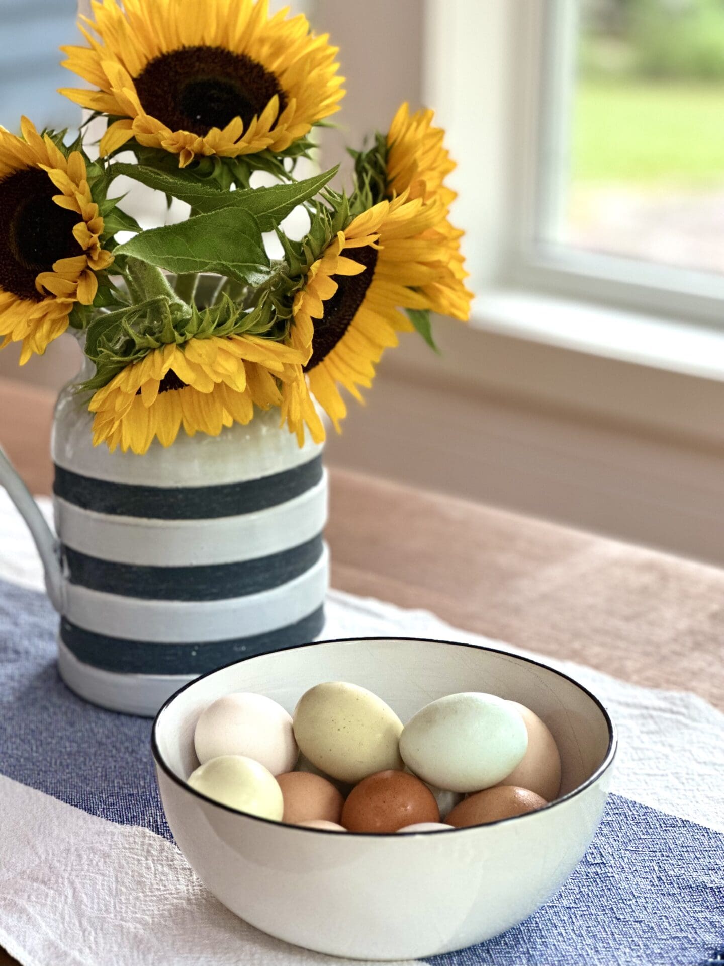 A bowl of pasted coloured eggs sits on a cottage kitchen table. A pitcher of yellow sunflowers is seen in the background.