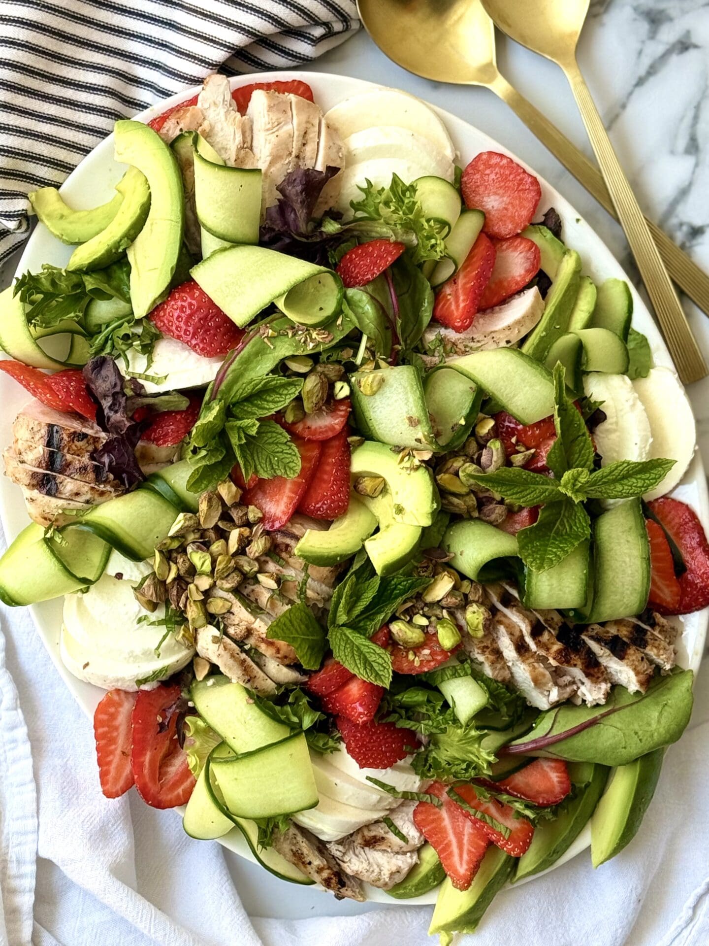 A large platter filled with Strawberry Cucumber Salad with Grilled Lemon Chicken is seen from above. The salad is beautifully arranged to show off its cucumber curls, bright red berries, creamy chunks of fresh mozzarella cheese and sprinkling of crunchy pistachios. A pair of gold-coloured salad servers sits nearby.