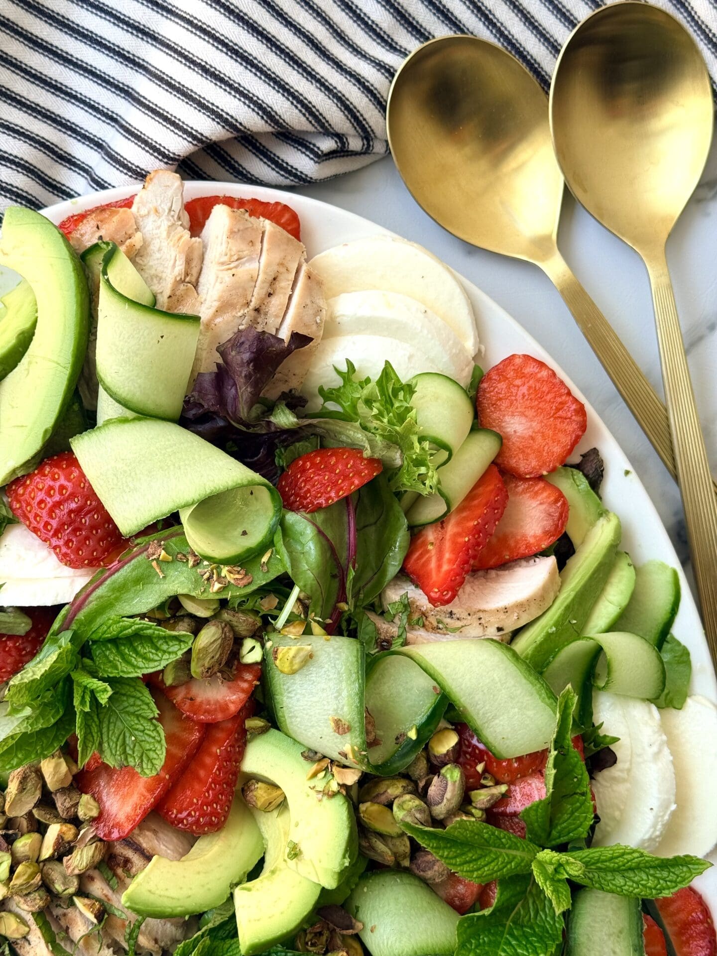 IMG_8875 A platter full of Strawberry Cucumber Salad with Grilled Lemon Chicken is seen from above, showcasing its beautiful colours and fresh fruits and vegetables. A set of gold salad servers and a black and white striped napkin complete the photo.