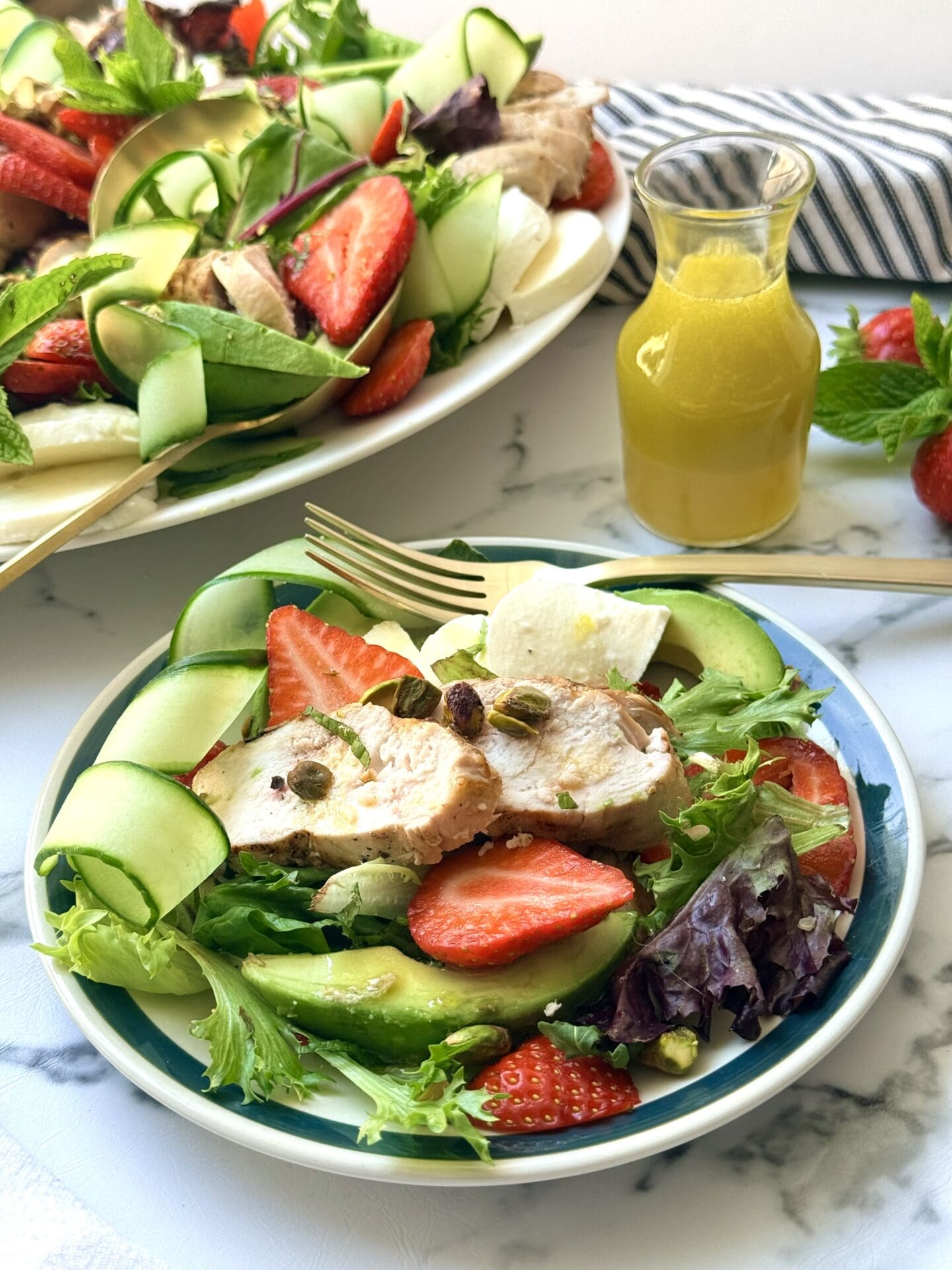 IMG_8890 A plate of Strawberry Cucumber Salad with Grilled Lemon Chicken sits on a white marble counter, waiting to be eaten with a gold fork. The platter of salad and bottle of lemon dressing are seen in the background.
