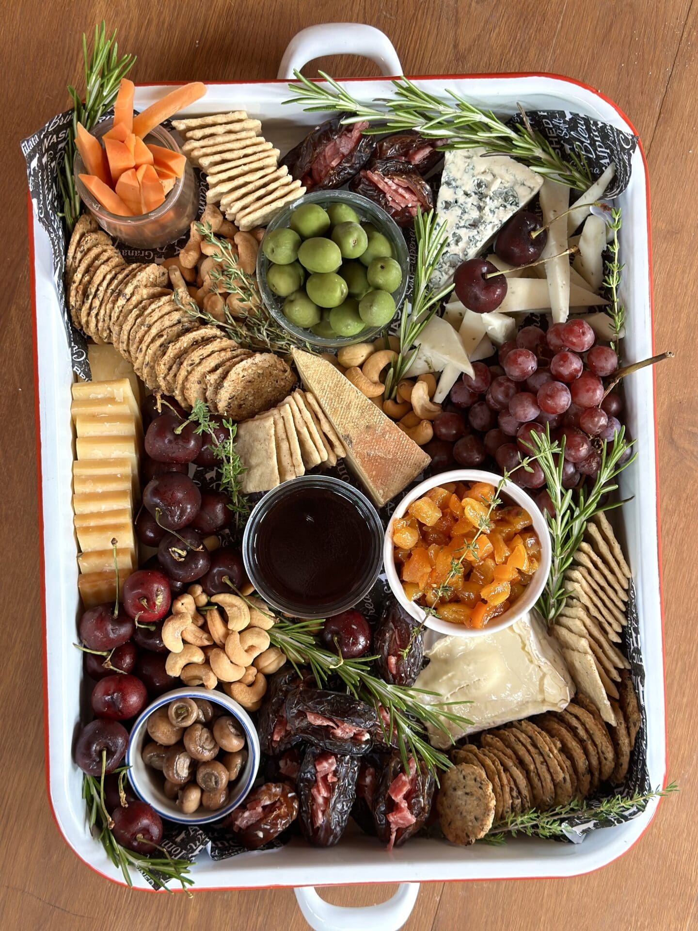 A enamelled metal tray is seen from above, filled with a lavish appetizer spread of meats, cheeses, olives, fresh fruit and pickles.
