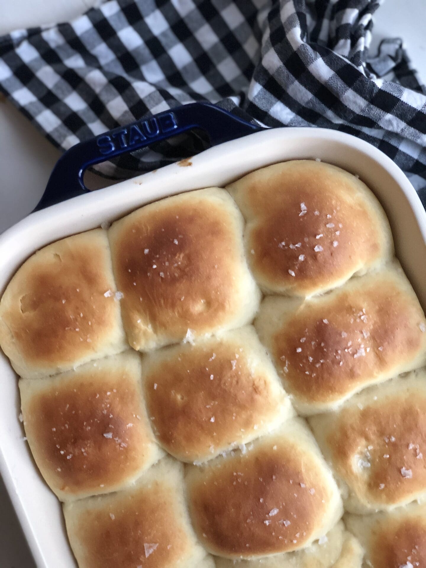 A pan of fresh-from-the-oven dinner rolls is seen from above, nestled together. The tops of the buns are golden brown and sprinkled with flaky sea salt.
