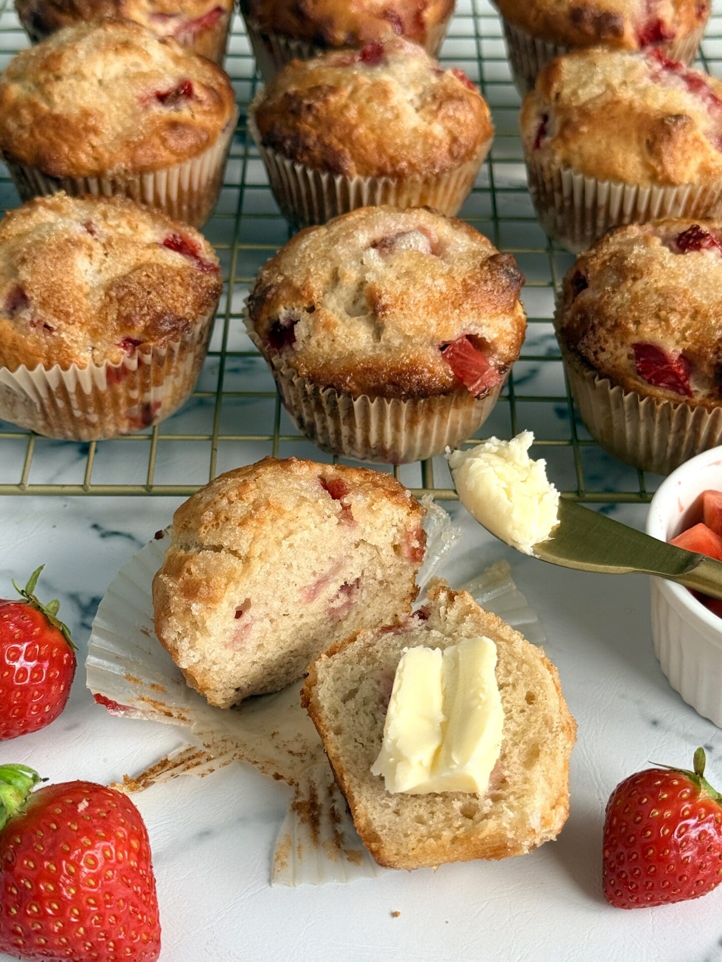 A strawberry yogurt muffin sits in the foreground, split in half and slathered in butter. The rest of the batch of muffins cools on a rack in the background. Juicy red strawberries decorate the scene.