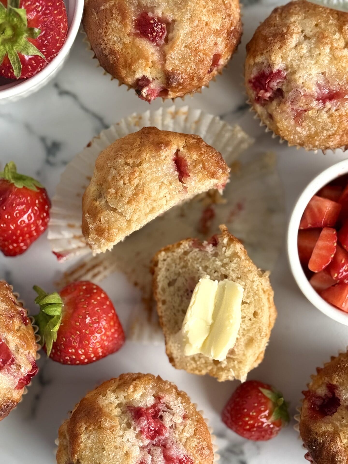 IMG_8940 A strawberry yogurt muffin is seen from above, split in two and slathered in butter. Fresh strawberries and more golden-topped muffins complete the scene.