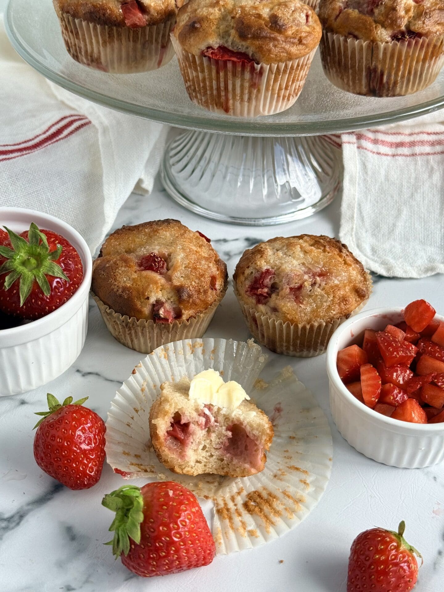 IMG_8950 A large bite has been taken out of a freshly baked, butter-topped, strawberry yogurt muffin, revealing its strawberry studded interior. A glass cake plate filled with muffins and bowls of fresh strawberries are seen in the background.