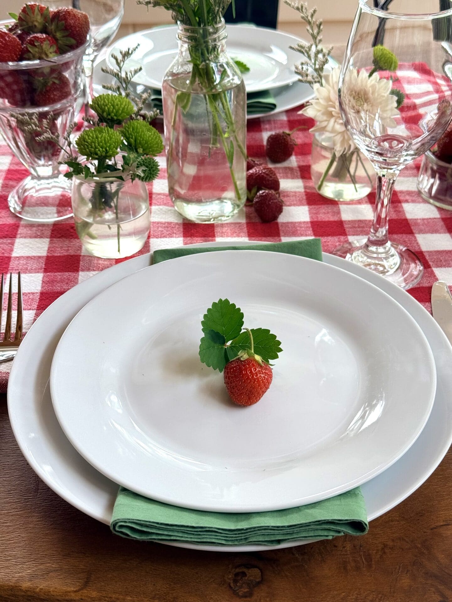 IMG_9492 An individual place setting at a Strawberry Social lunch is the focus of this photo. The place setting features white plates, a green cloth napkin, stemware and cutlery. The plate is finished with a plump strawberry with strawberry leaves. The place setting rests on a red gingham tablecloth. Small vases of white and green wild flowers are seen in the background.