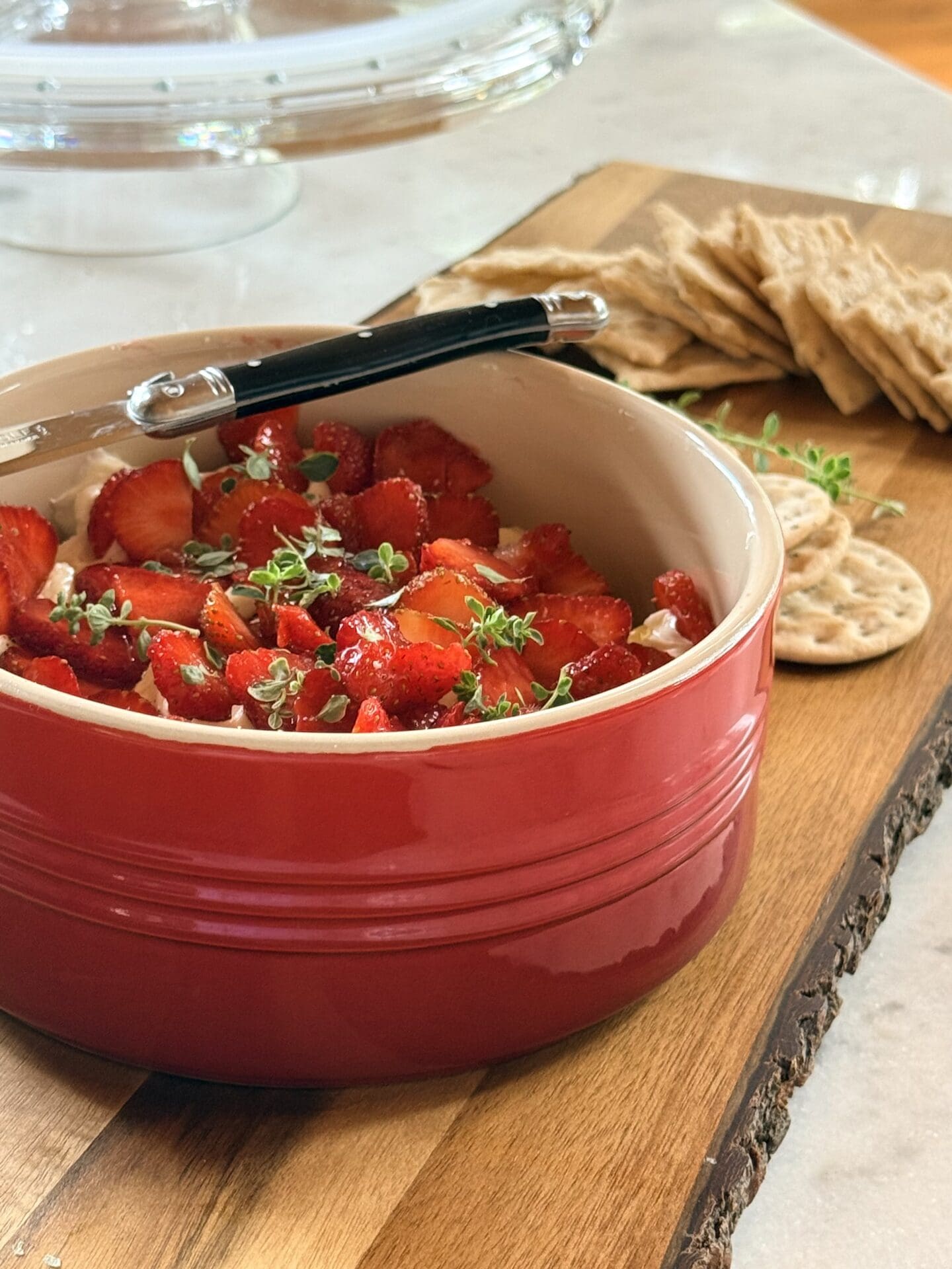A red bowl filled with Whipped Brie with Strawberries and Fresh Thyme sits on a wood board surrounded by crackers.