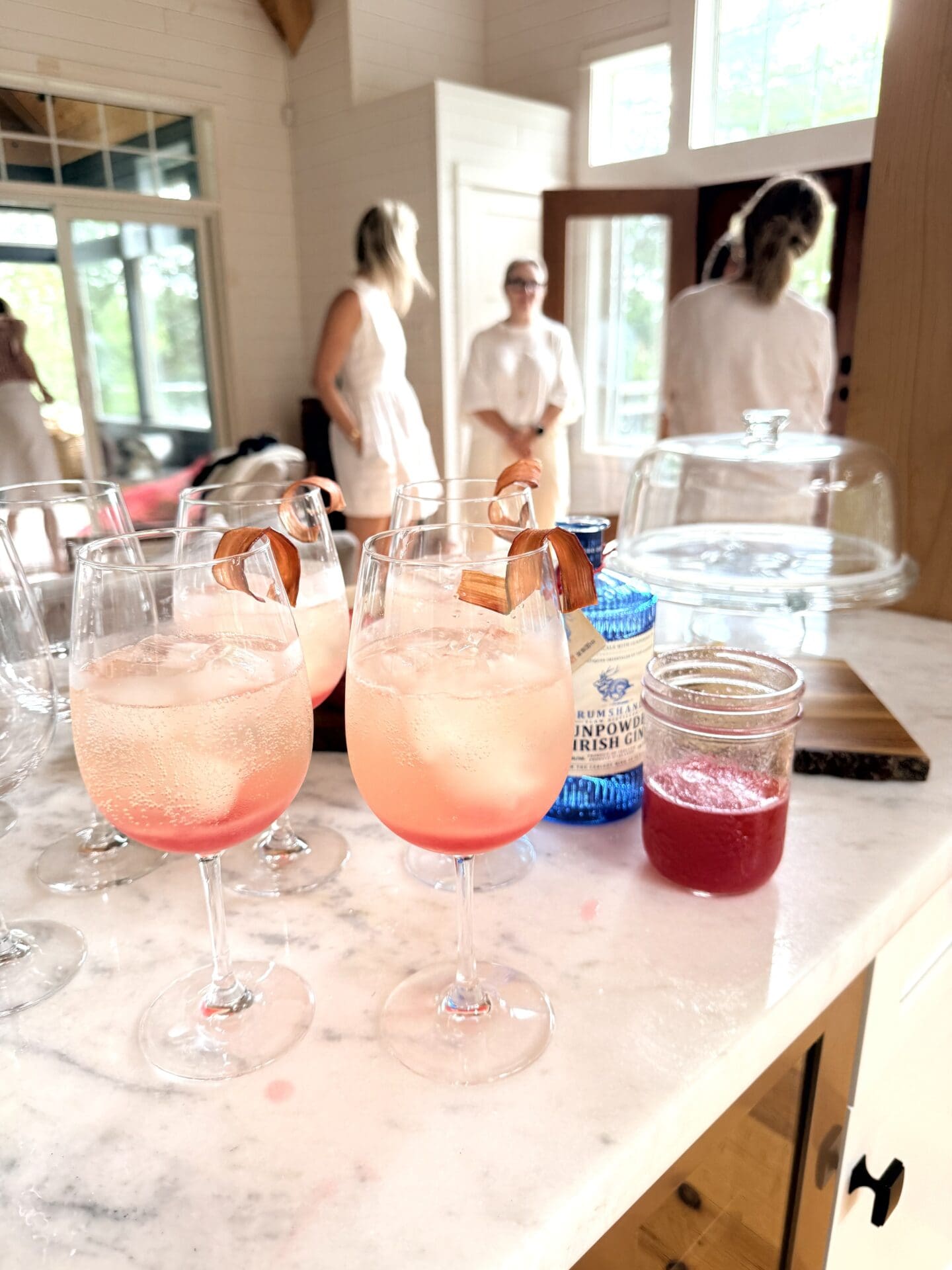 IMG_9507 A group of Rhubarb Gin Spritz Cocktails sit on a marble bar. Party guests are seen in conversation in the background.