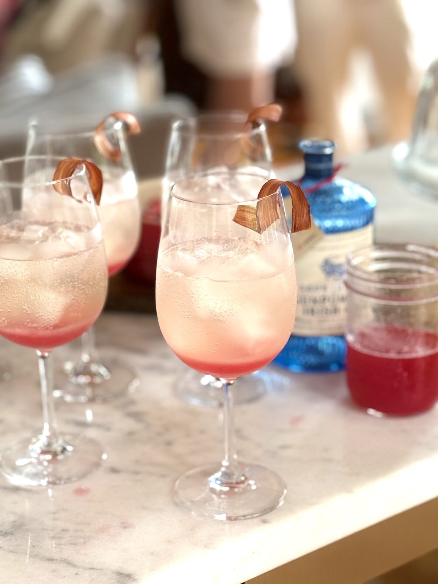 Four Rhubarb Gin Spritz cocktails are being made on a white marble bar. A blue bottle of gin is seen in the background, along with a jar of ruby red rhubarb syrup.