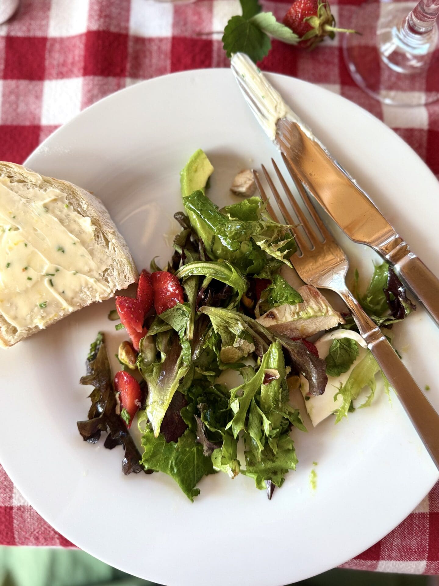 A dinner plate containing salad is seen from above. A slice of bread, slathered in lemon chive butter perches on the edge of the plate. The plate rests on a red gingham tablecloth.