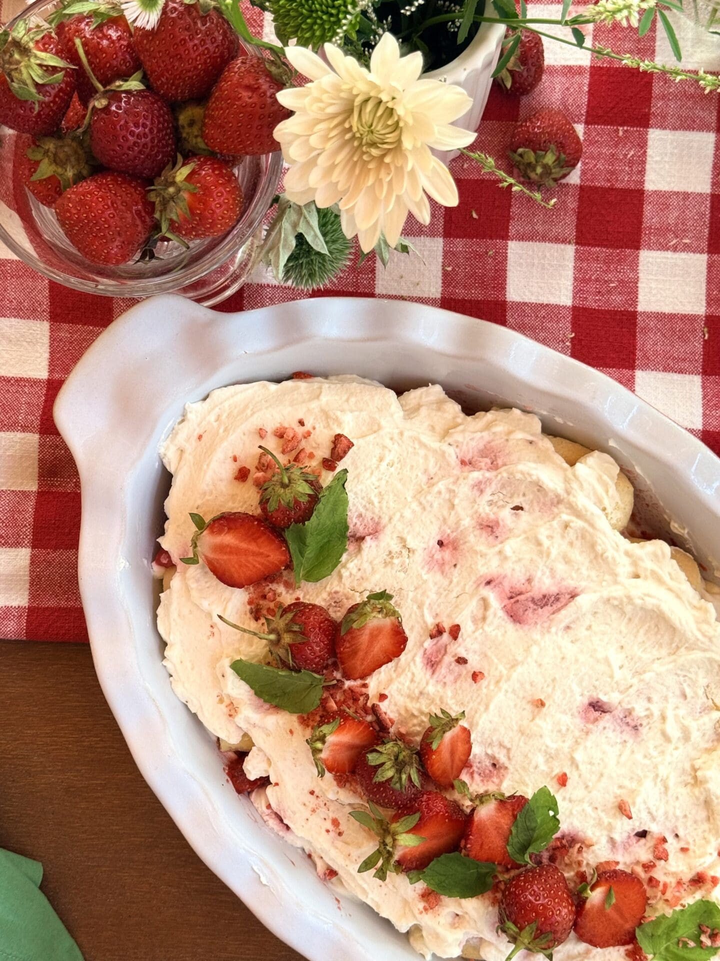 IMG_9548 A large serving dish of strawverry tiramisu is seen from above, resting on a red and white tablecloth. The tiramisu is beautifully garnished with fresh strawberries and mint.