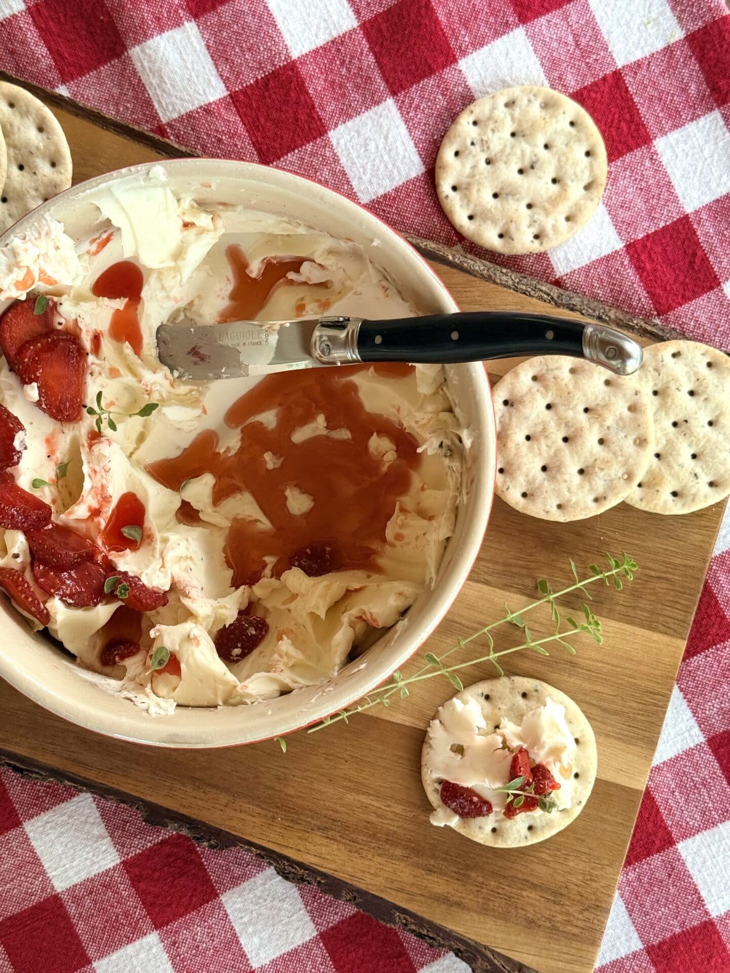 A bowl of whipped Brie with stawberries and thyme is seen from above on a red gingham tablecloth. The appetizer is being devoured, showing the swoops of whipped cheese under the layer of fresh strawberries. A cracker with a generous amount of the whipped Brie sits beside the bowl, ready to be eaten.