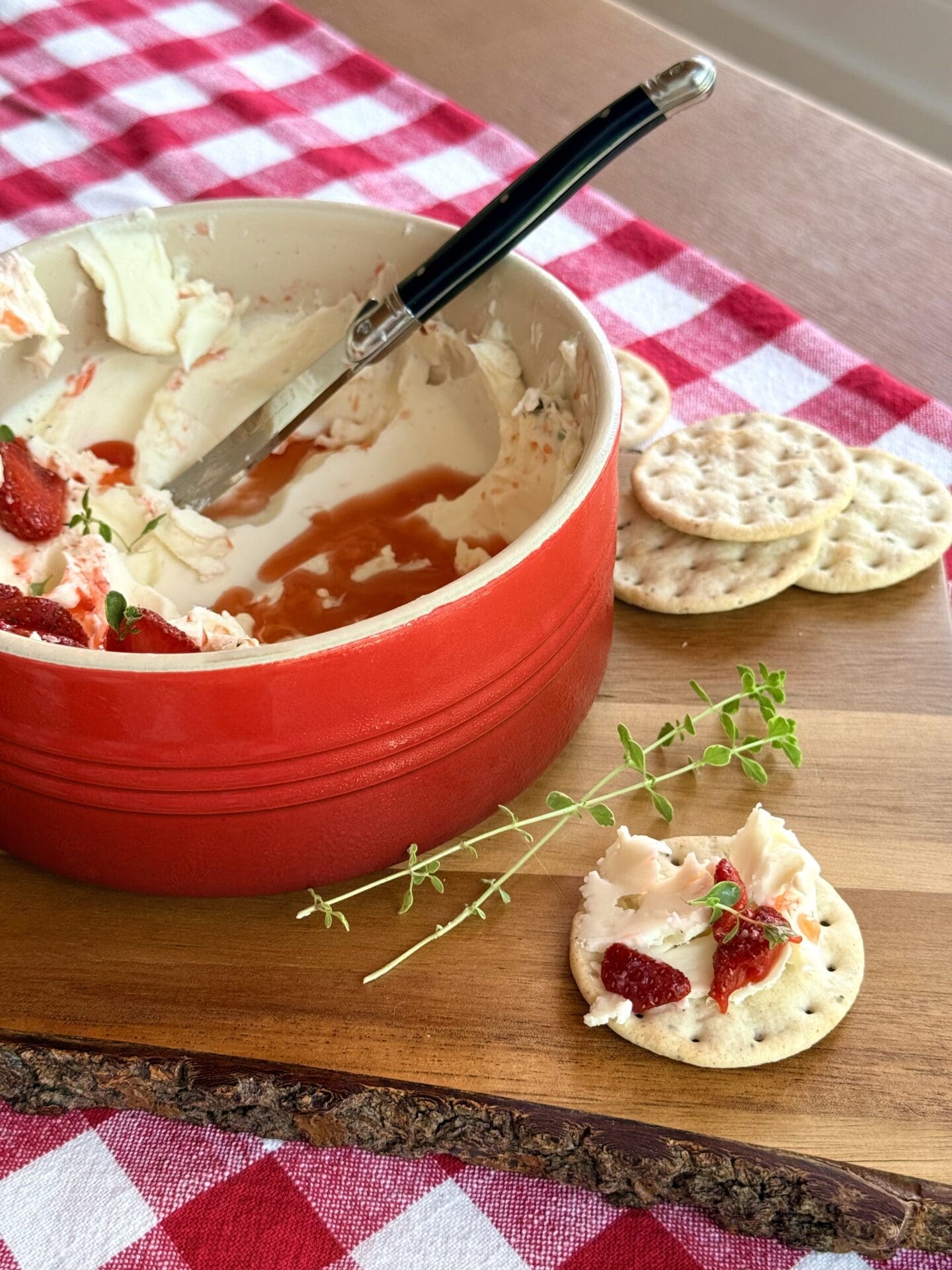 A bowl of Whipped Brie with Strawberries and Fresh Thyme sits on a wood board. The appetizer has been partially enjoyed, showing the swoops of creamy cheese and juicy strawberry topping. A cracker sits in the foreground, topped with the brie and strawberry mixture.