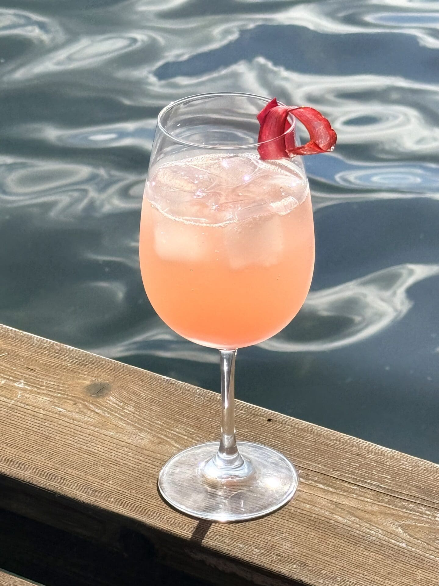 A large stemmed glass is filled with an ice-cold, blush pink, Rhubarb Gin Spritz cocktail. The drink rests on a wood railing, overlooking a sparkling lake.