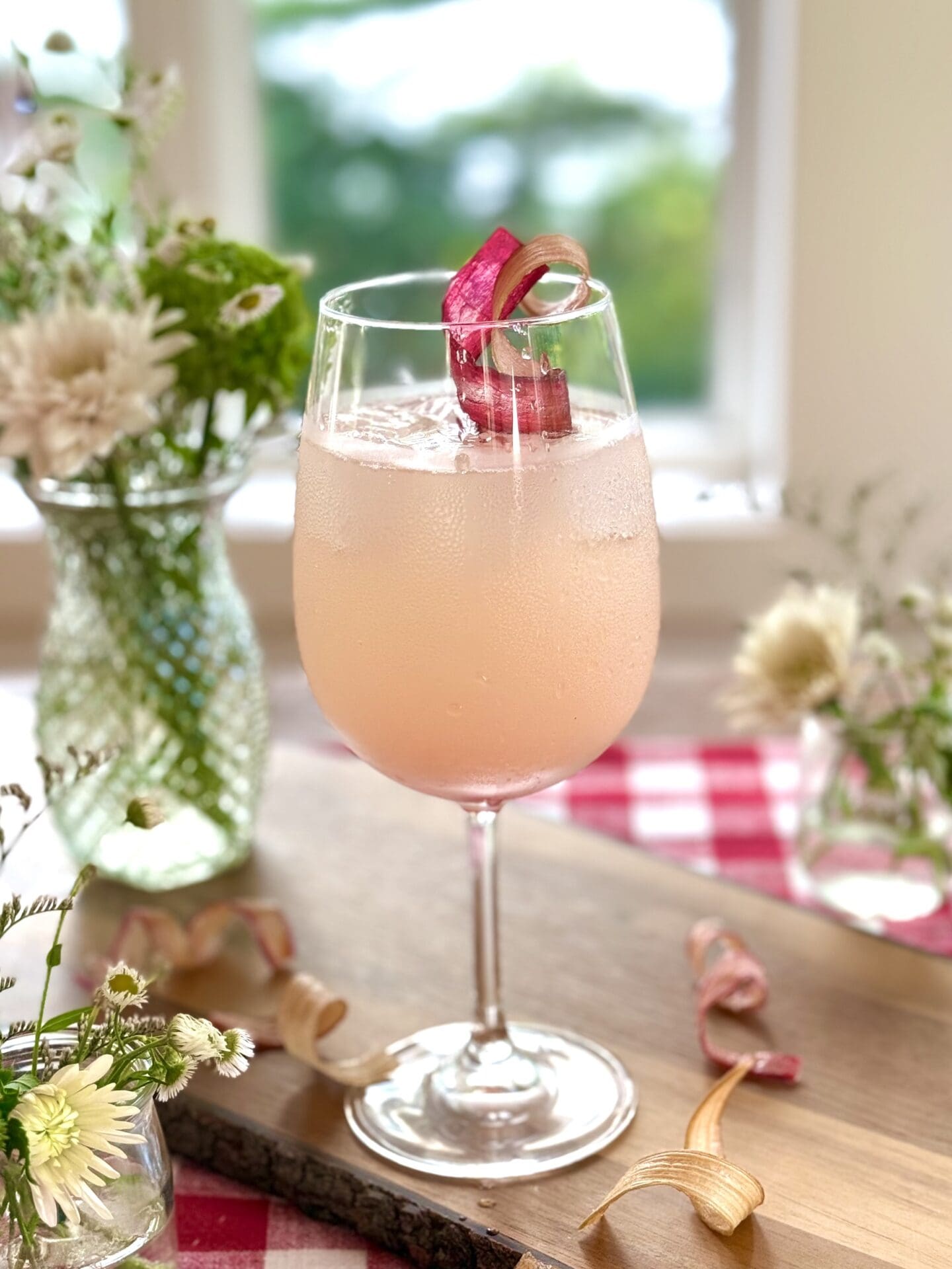 A rhubarb gin spritz cocktail sits on a wood board in front of a window. A summery scene of trees and a lake can be seen through the window. The drink is surrounded by bouquets of wildflowers. A red gingham tablecloth is seen in the background.