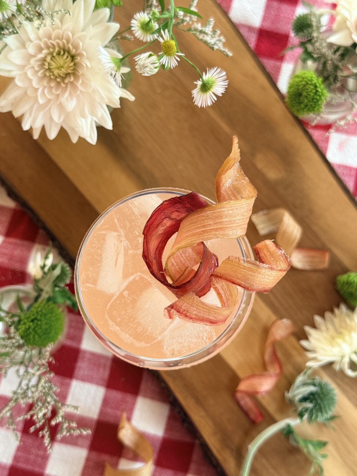 A cocktail glass is seen from above, filled with an ice cold pink drink and garnished lavishly with candied rhubarb ribbons. The glass sits on a wood board, surrounded by flowers. A red gingham tablecloth completes the image.