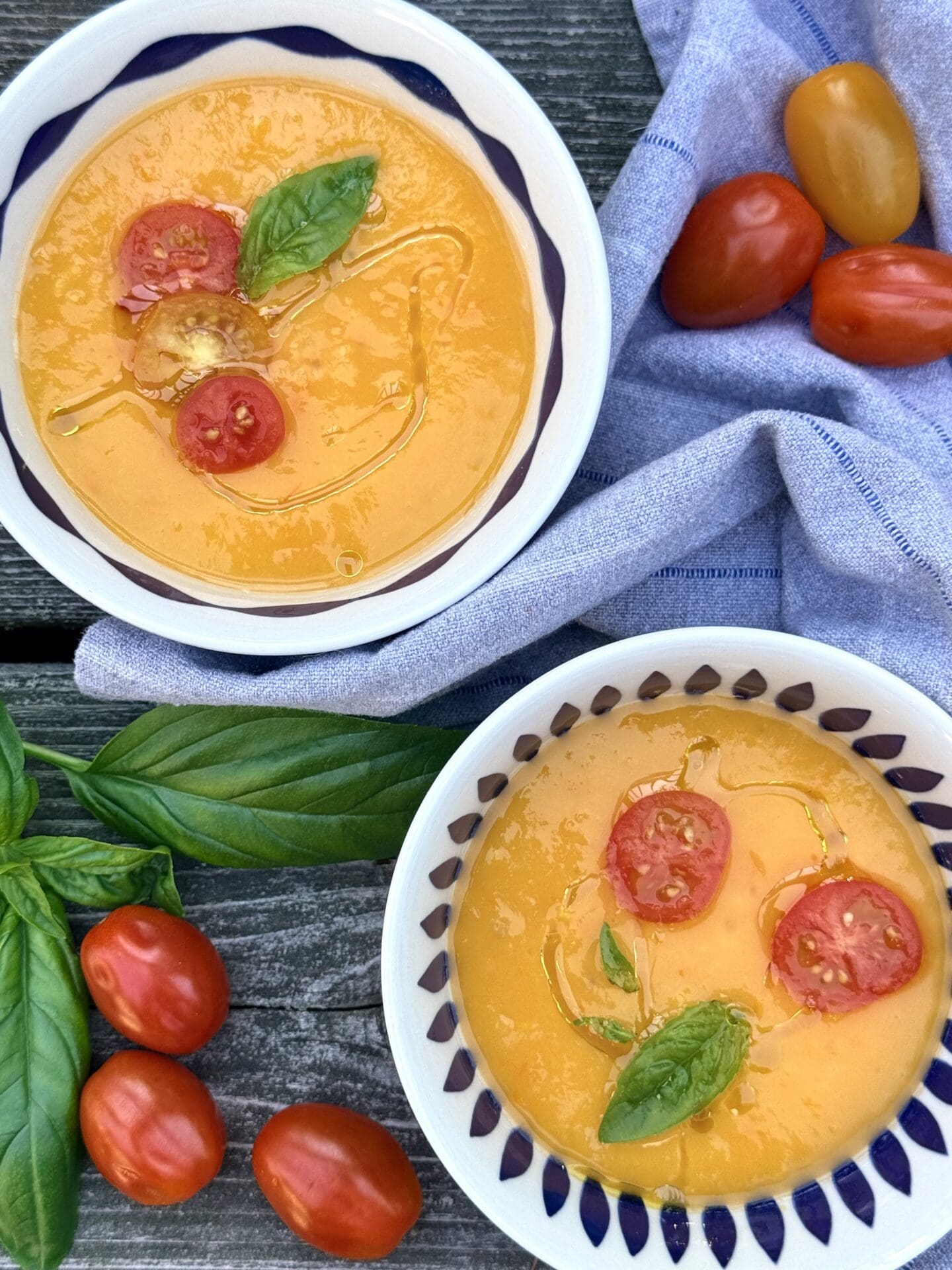 Two bowls of Yellow Tomato Gazpacho are seen from above, showcasing its beautiful sunny yellow colour. The bowls of soup are garnished with sliced cherry tomatoes and basil leaves.